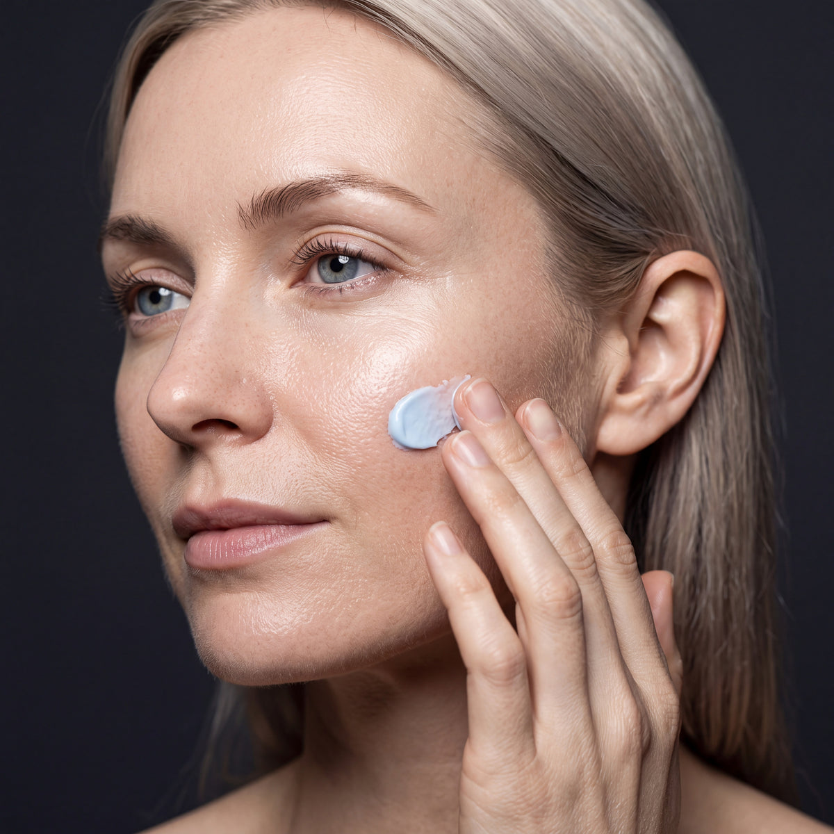 Woman applying a small dab of pale blue cream to her cheekbone with her fingertips