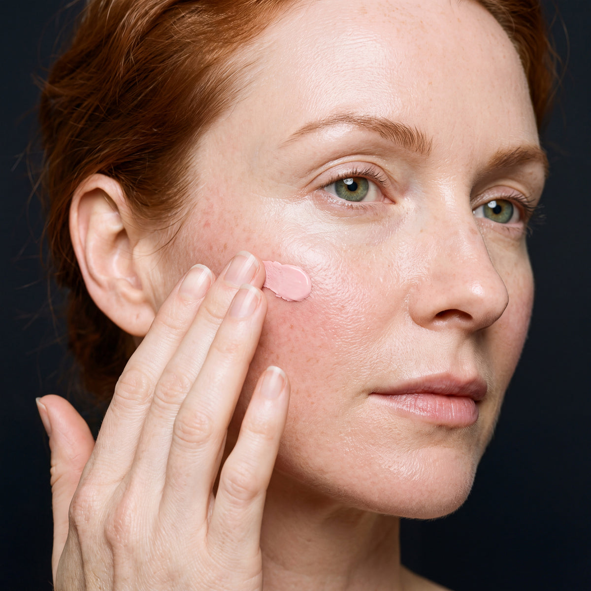 Woman applying a small dab of pale pink cream to her cheekbone with her fingertips