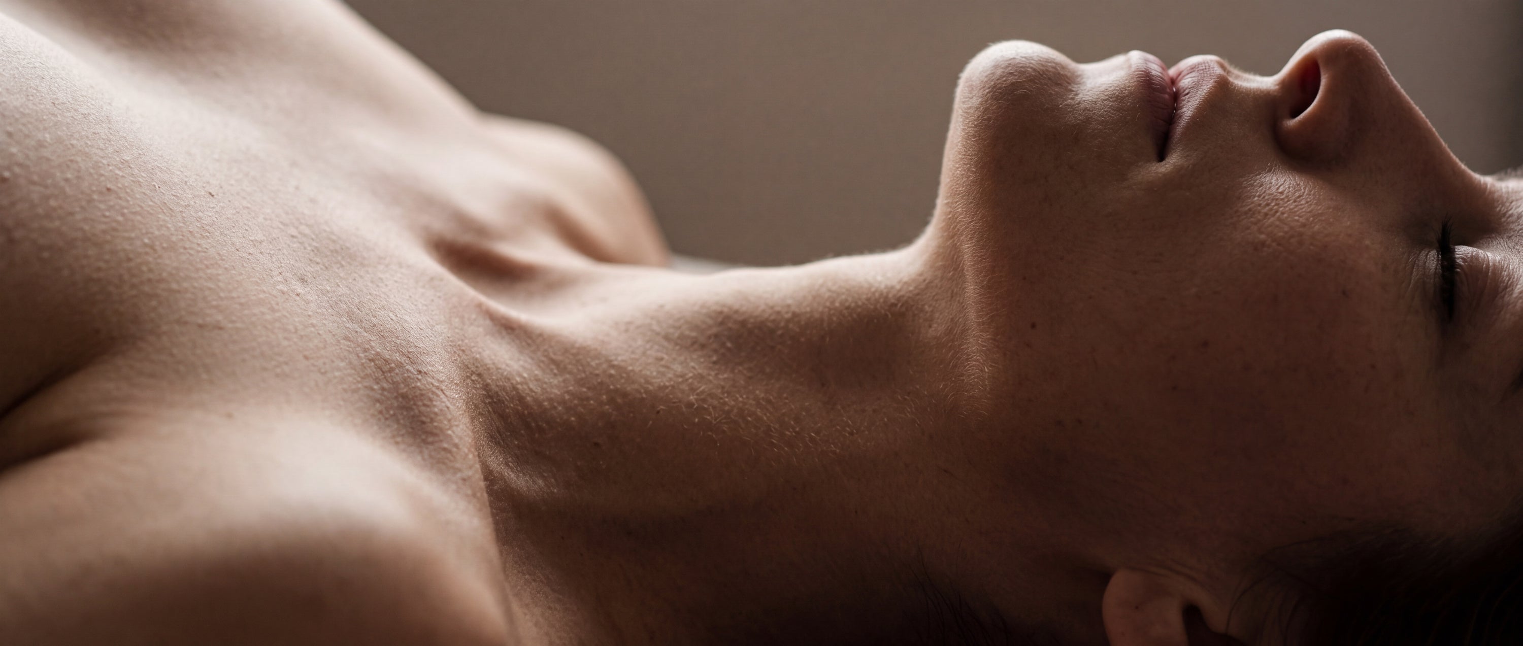 Close-up of a reclining woman's shoulder, throat and jaw lit softly against a warm neutral backdrop