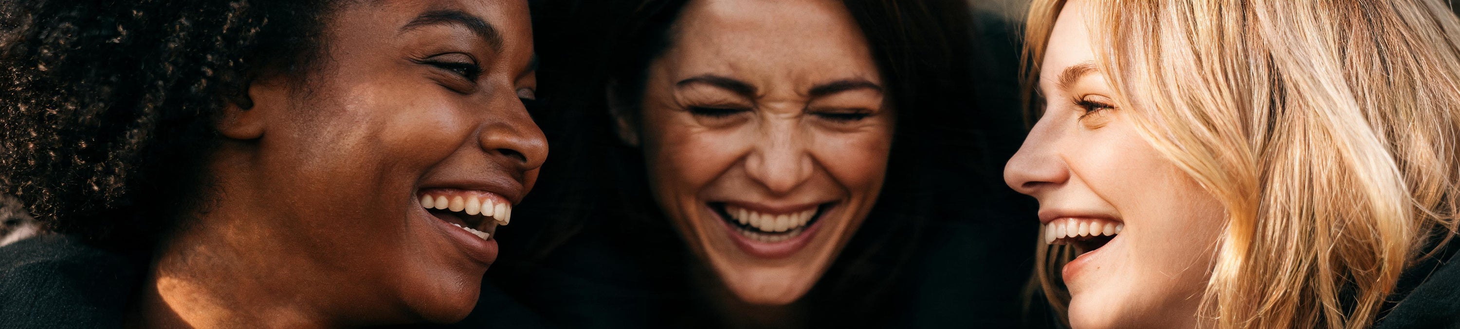 Three women laughing together in close-up, lit warmly against a dark background