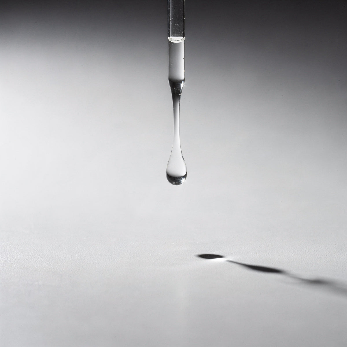 A glass pipette releasing a single clear droplet onto a pale grey surface