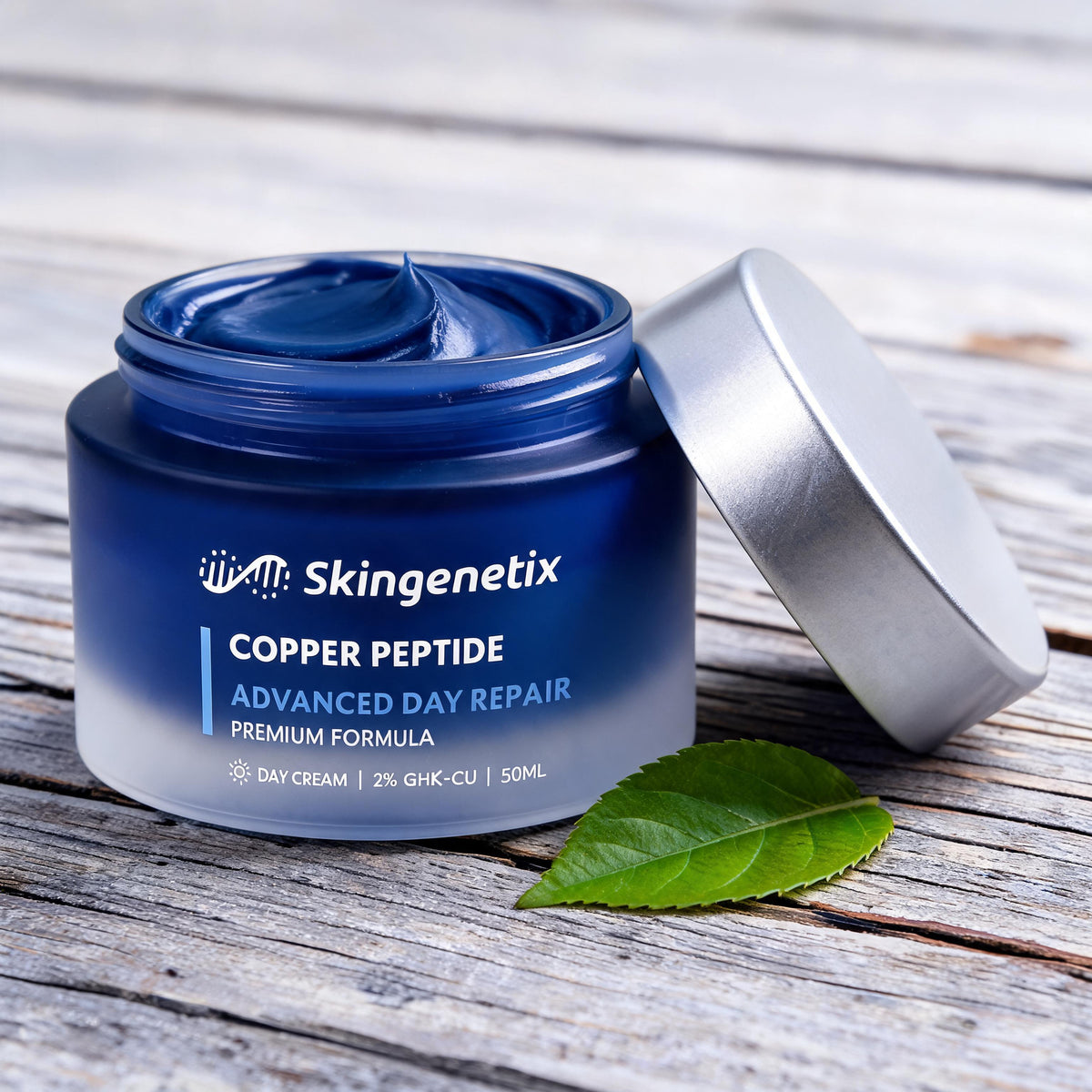 Open frosted navy glass jar of Skingenetix Copper Peptide Advanced Day Repair gel-cream, 50ml, on weathered pale wood with the silver lid propped beside it and a single green leaf in front
