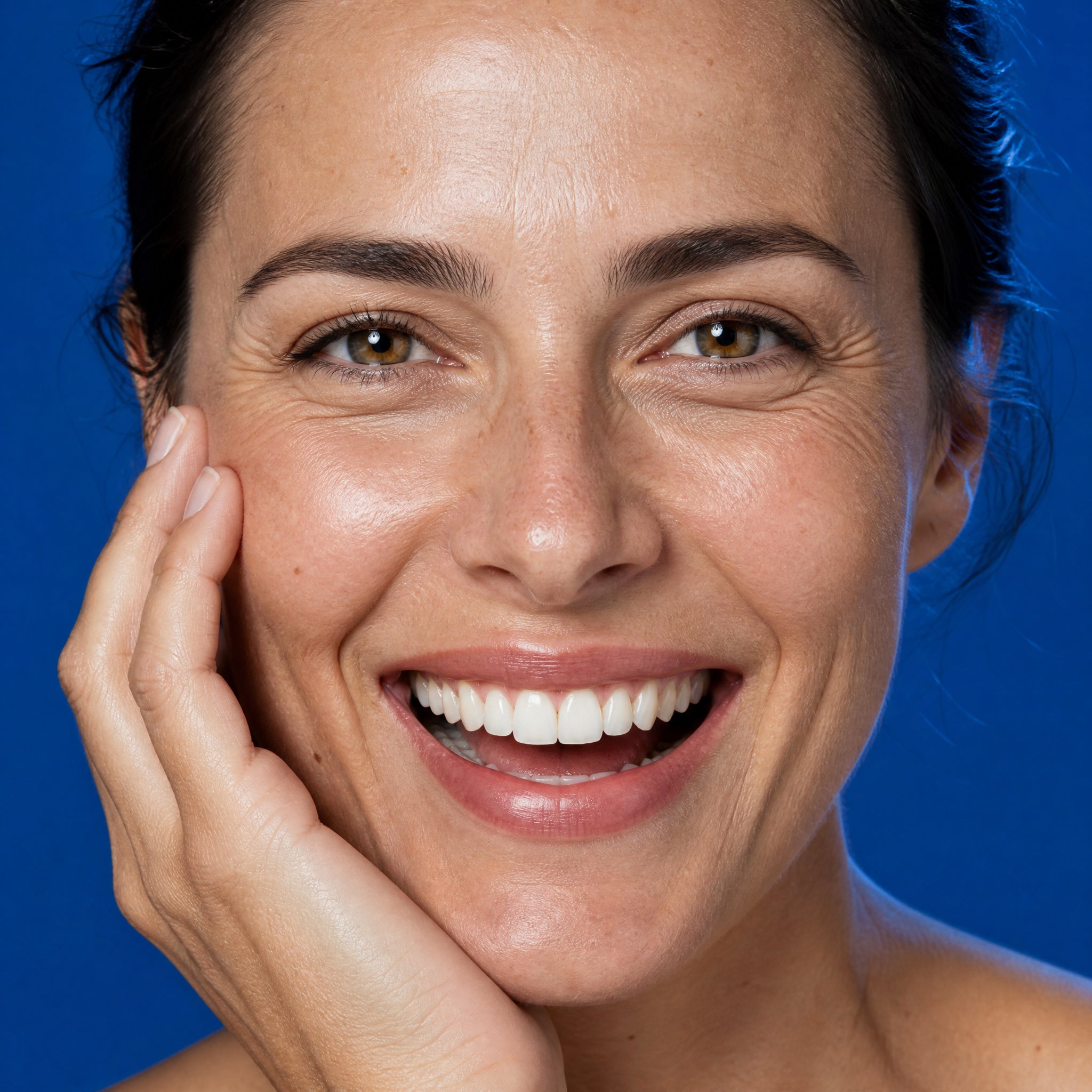 A smiling woman in her thirties resting her hand against her cheek on a deep blue background - Skingenetix copper peptide GHK-Cu skin appearance research