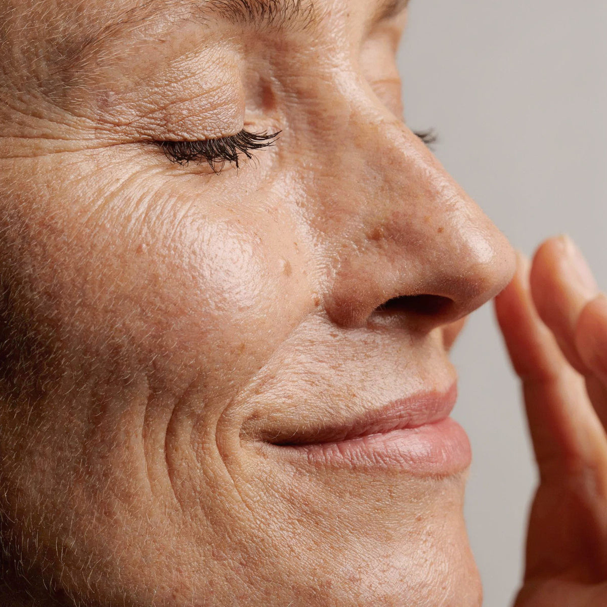 Close-up of a mature woman's face in near profile, eyes closed with a small smile, her fingertips resting lightly against her cheek, with crow's feet at the eye corner and fine lines around the mouth