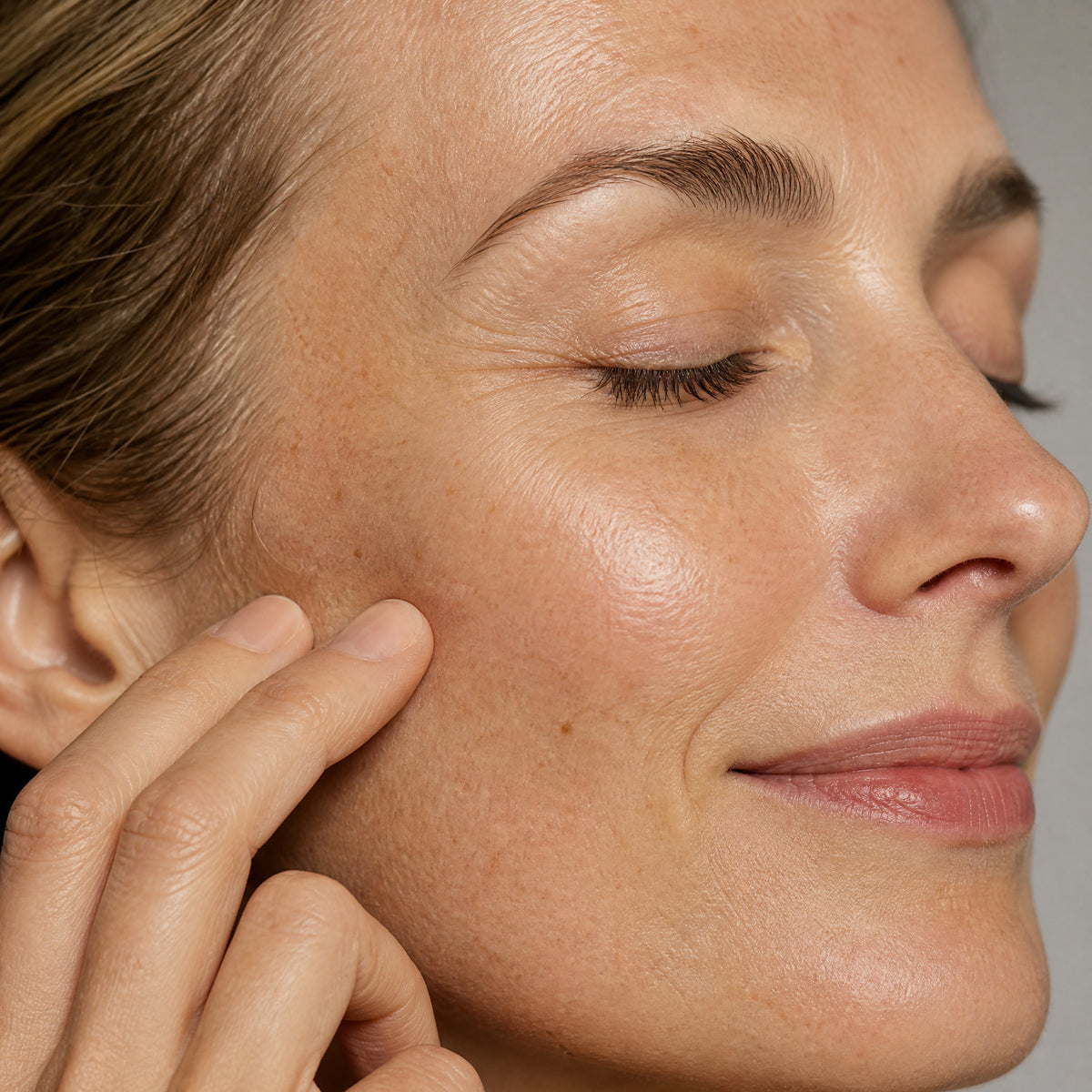 Close-up of a woman's face in near profile, eyes closed with a faint smile, the backs of her fingers resting against her cheek and jawline