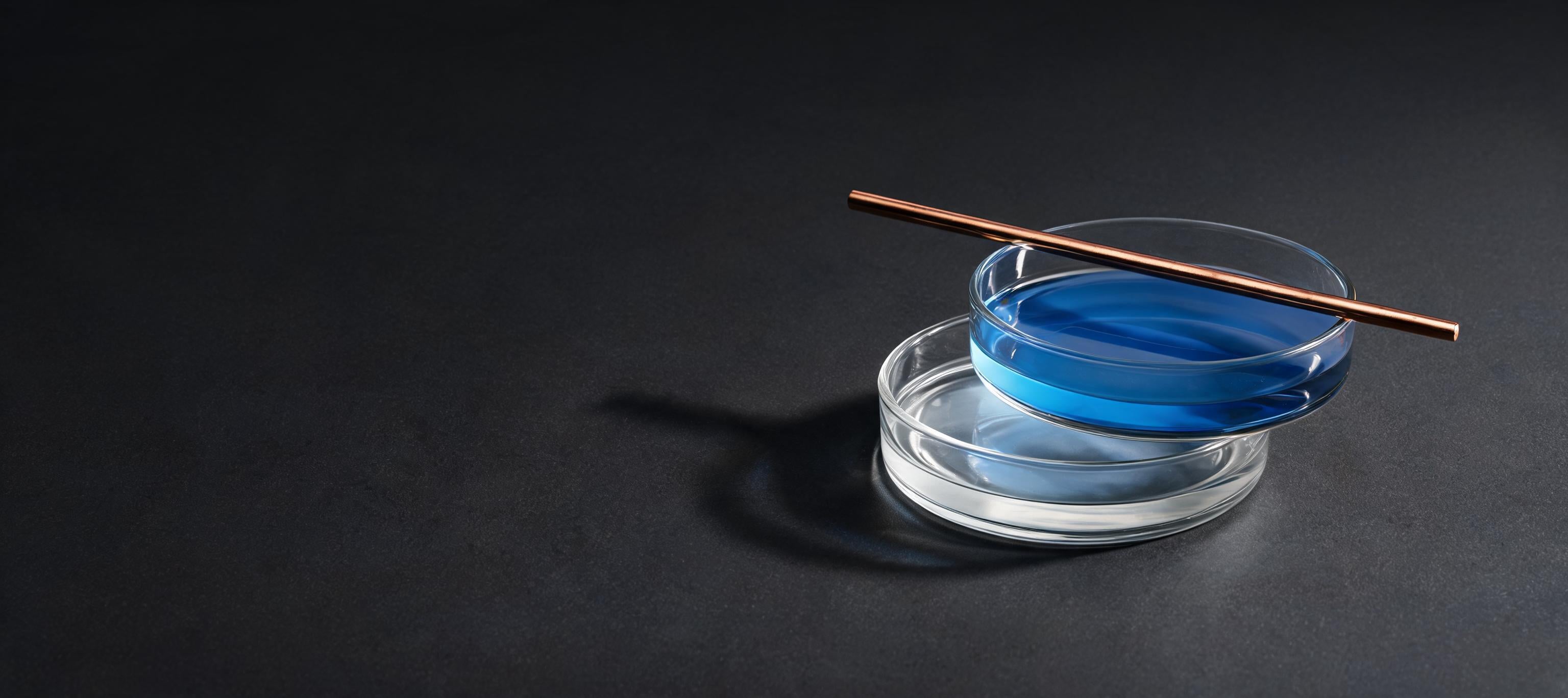Two stacked laboratory glass dishes holding clear serum and a blue peptide solution with a copper rod resting across them, lit against a graphite background