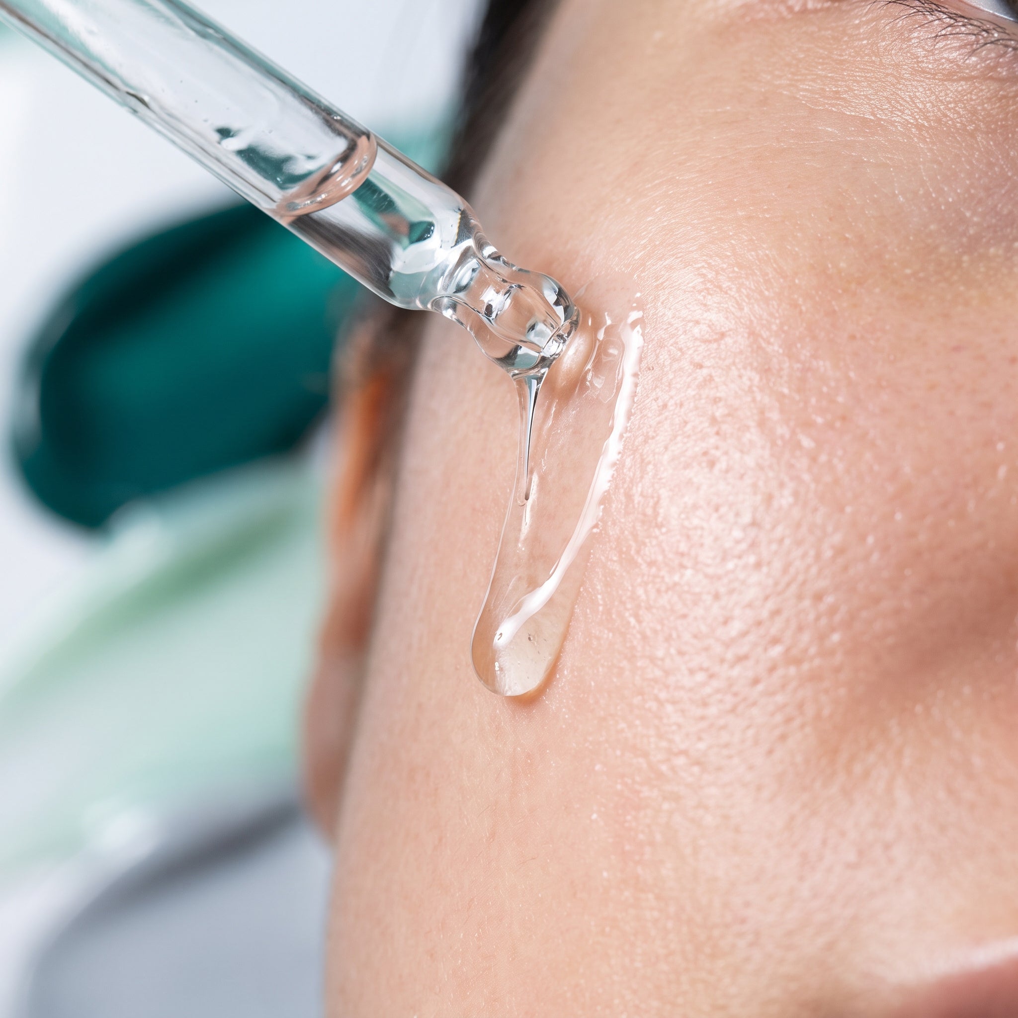 Glass dropper releasing Matrixyl 3000 firming serum onto a woman's cheekbone, the serum running down her skin in close-up