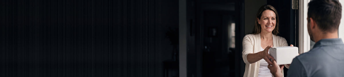 A woman smiling as she takes a plain white parcel from a delivery driver at her door