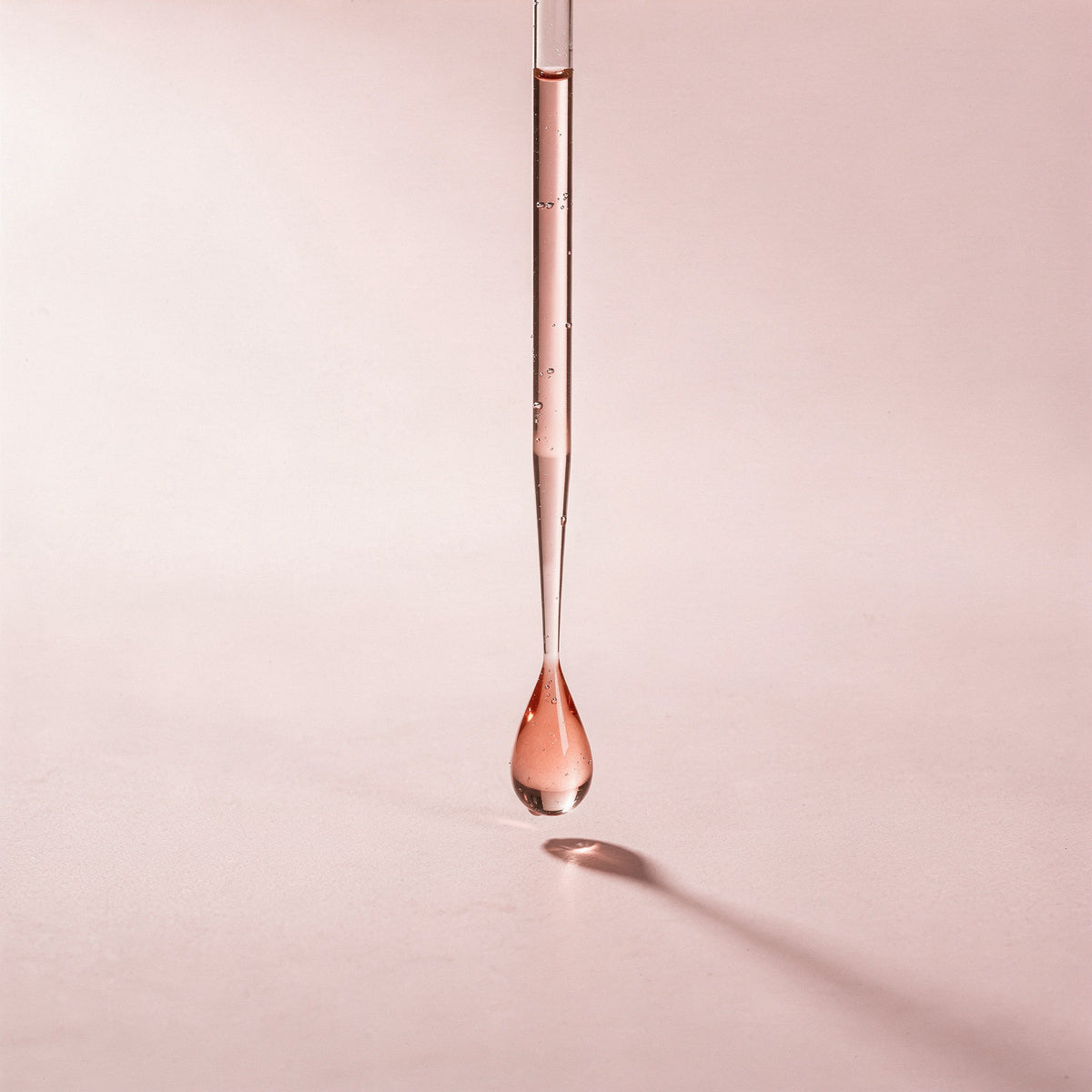 A glass pipette releasing a single pink droplet onto a rose-coloured surface