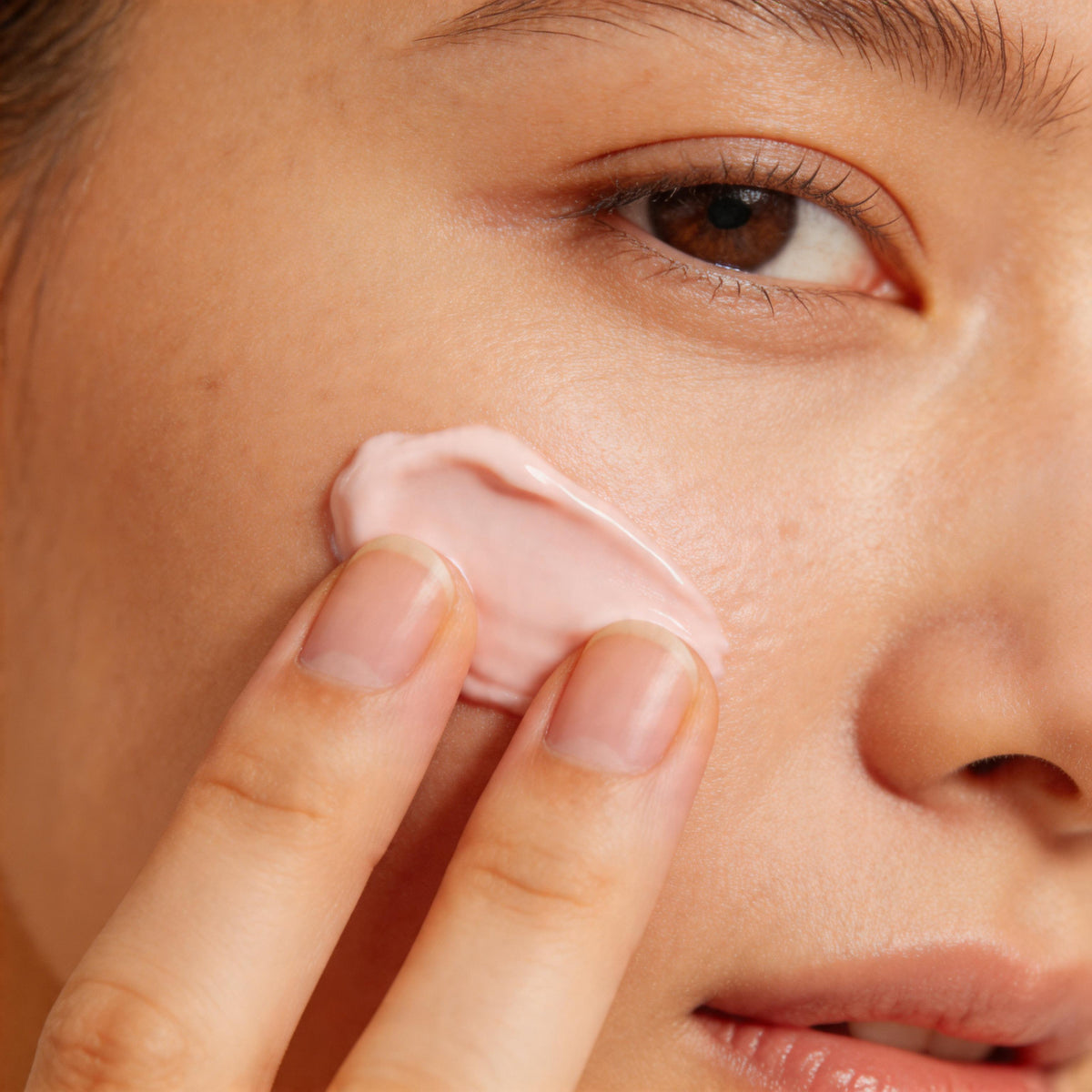 Two fingertips smoothing a soft pink cream onto the cheekbone in close-up, illustrating the skin renewal PDRN is studied for